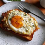 A perfectly fried egg with a runny yolk sits atop crispy feta cheese and toast, seasoned with herbs and pepper.
