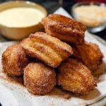 A pile of golden-brown Easy Baked Churro Bites coated in cinnamon sugar, with a bowl of dipping sauce in the background.