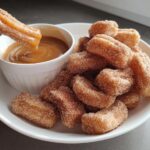 A hand dips a fluffy air fryer churro bite into a bowl of caramel sauce, with a pile of churro bites on a plate.