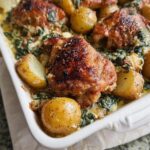 Close-up of a white baking dish filled with golden brown Garlic Parmesan Chicken Thighs and Potatoes nestled in a creamy spinach sauce.