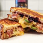 A gourmet blueberry grilled cheese sandwich, cut in half, revealing melted cheese, bacon, and jalapenos.