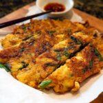 A close-up of freshly made Korean Pancakes (Pajeon) sliced and served on a paper towel with chopsticks and a dipping sauce.