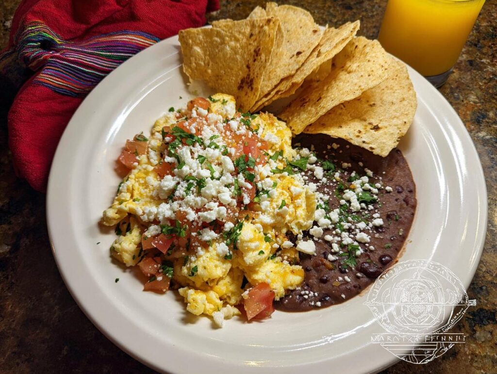A plate of scrambled Mexican Eggs topped with diced tomatoes and crumbled white cheese, served with refried beans and tortilla chips.