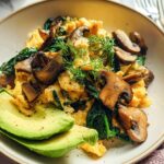 A close-up of delicious Mushroom Spinach Scrambled Eggs served with sliced avocado and fresh dill.