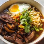 A delicious bowl of Slow Cooker Beef Ramen Noodles topped with a fried egg, sliced mushrooms, and fresh cilantro.