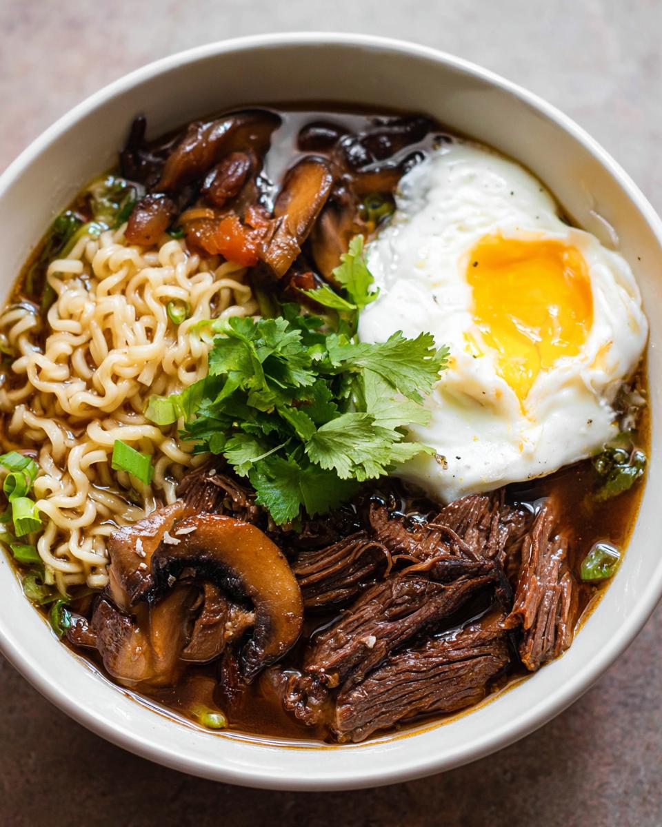A close-up of a bowl of Slow Cooker Beef Ramen Noodles, featuring tender beef, ramen noodles, mushrooms, a fried egg, and cilantro.