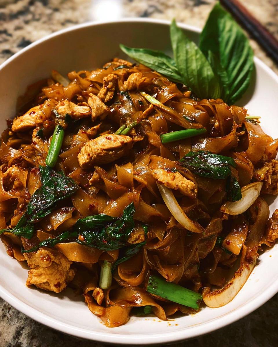 A close-up of a bowl of Street Style Thai Drunken Noodles with chicken, wide rice noodles, and fresh basil.