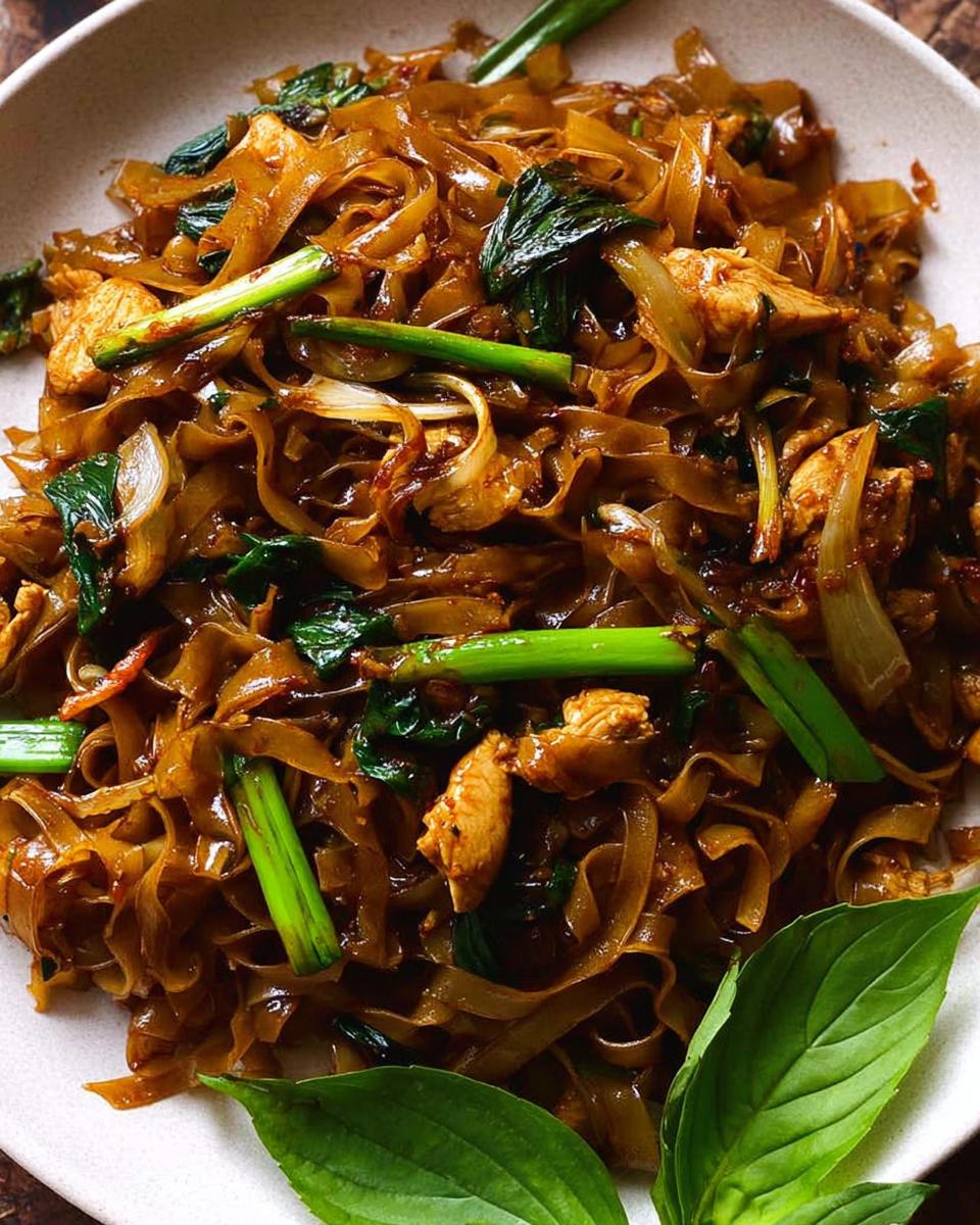 A close-up of a plate piled high with glossy, wide rice noodles, chicken pieces, green onions, and bok choy, representing Street Style Thai Drunken Noodles.