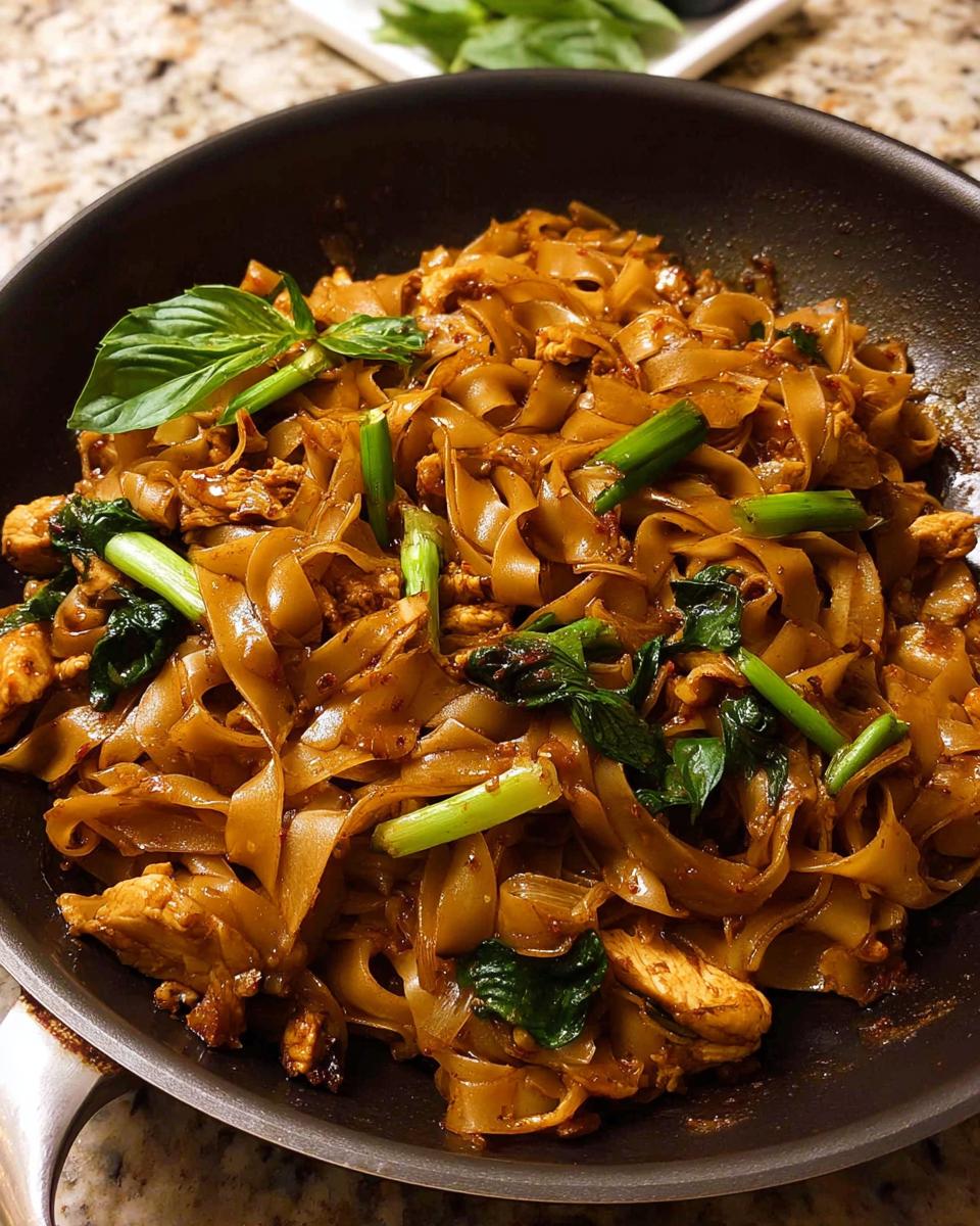 Close-up of Street Style Thai Drunken Noodles with chicken, green onions, and basil in a black pan.