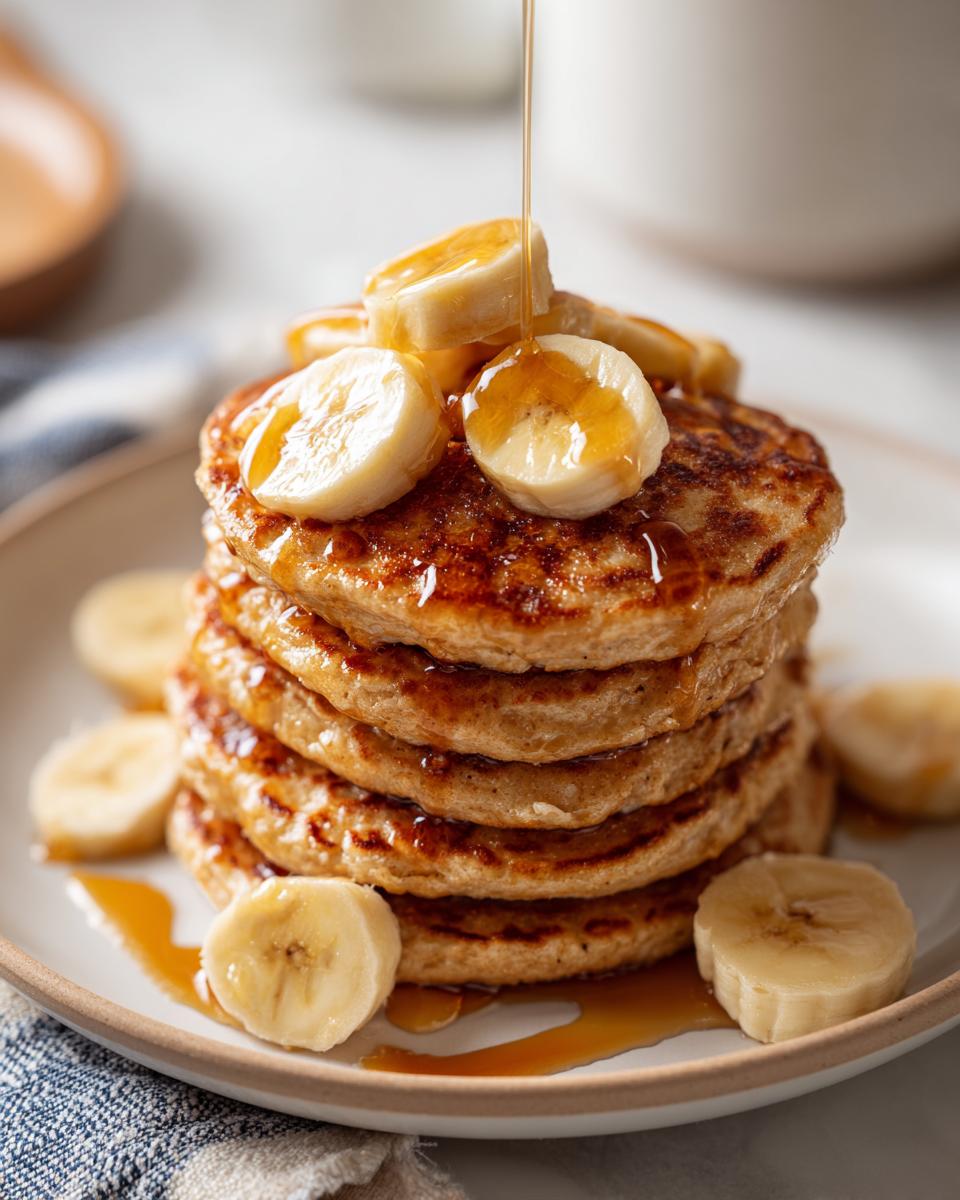 A tall stack of golden-brown banana oat pancakes, topped with fresh banana slices and drizzled with syrup.
