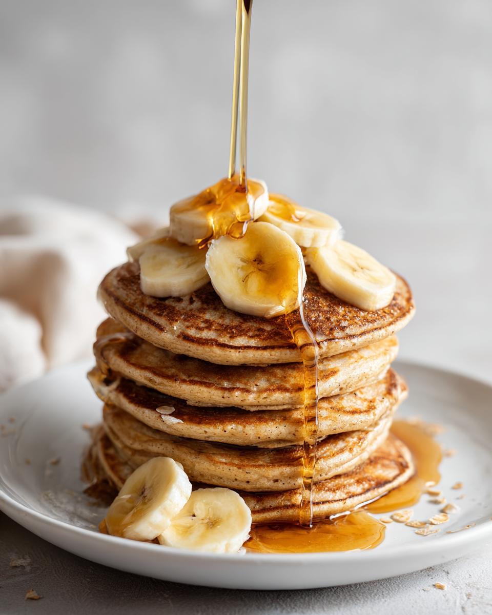 Stack of fluffy banana oat pancakes topped with sliced bananas and drizzled with syrup.