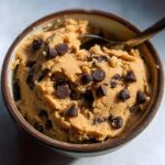 A bowl filled with delicious protein cookie dough, studded with chocolate chips, and a spoon resting on top.