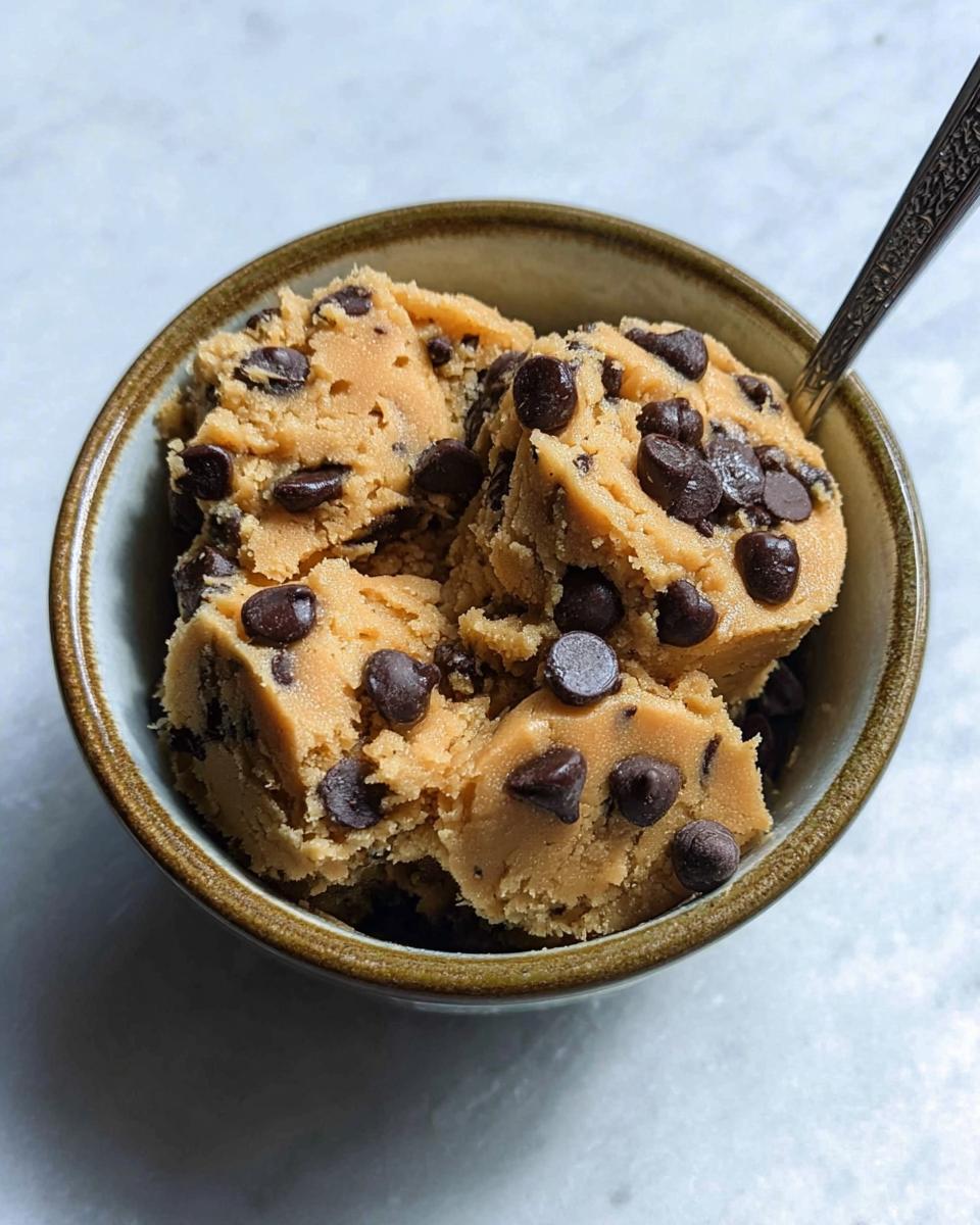 A bowl filled with delicious protein cookie dough studded with chocolate chips, with a spoon in the background.