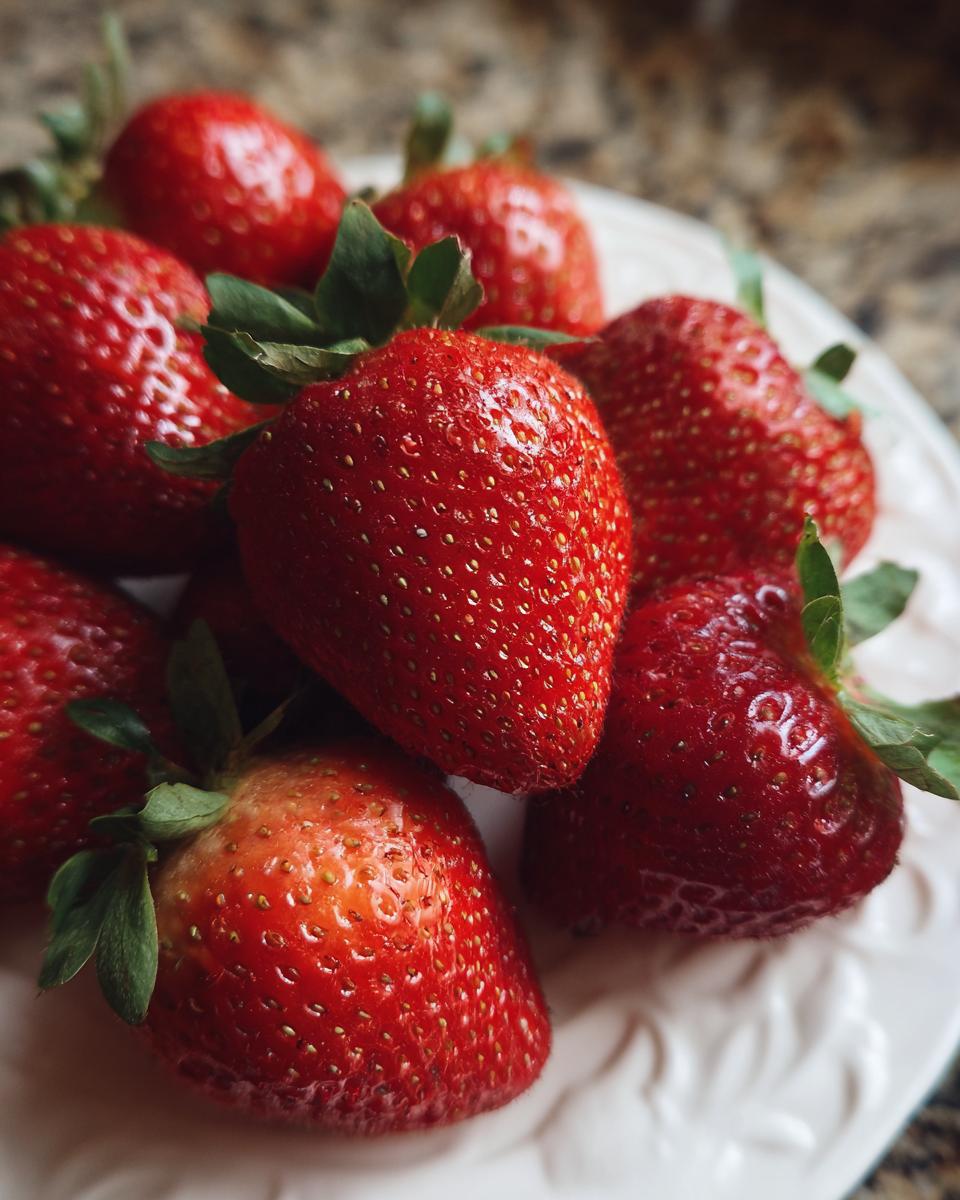 A close-up of fresh, ripe strawberries with green leaves on a white plate, perfect for strawberry spinach summer salad recipes.