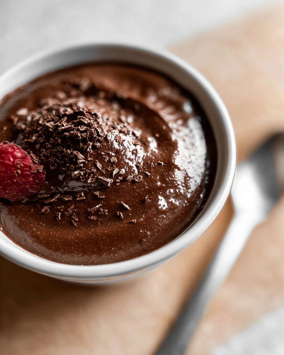 A bowl of decadent High Protein Chocolate Pudding topped with chocolate shavings and a fresh raspberry.
