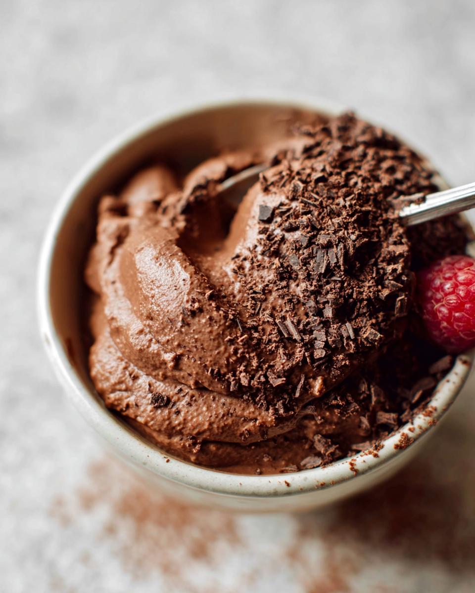 A close-up of a bowl of decadent High Protein Chocolate Pudding, topped with chocolate shavings and a fresh raspberry.