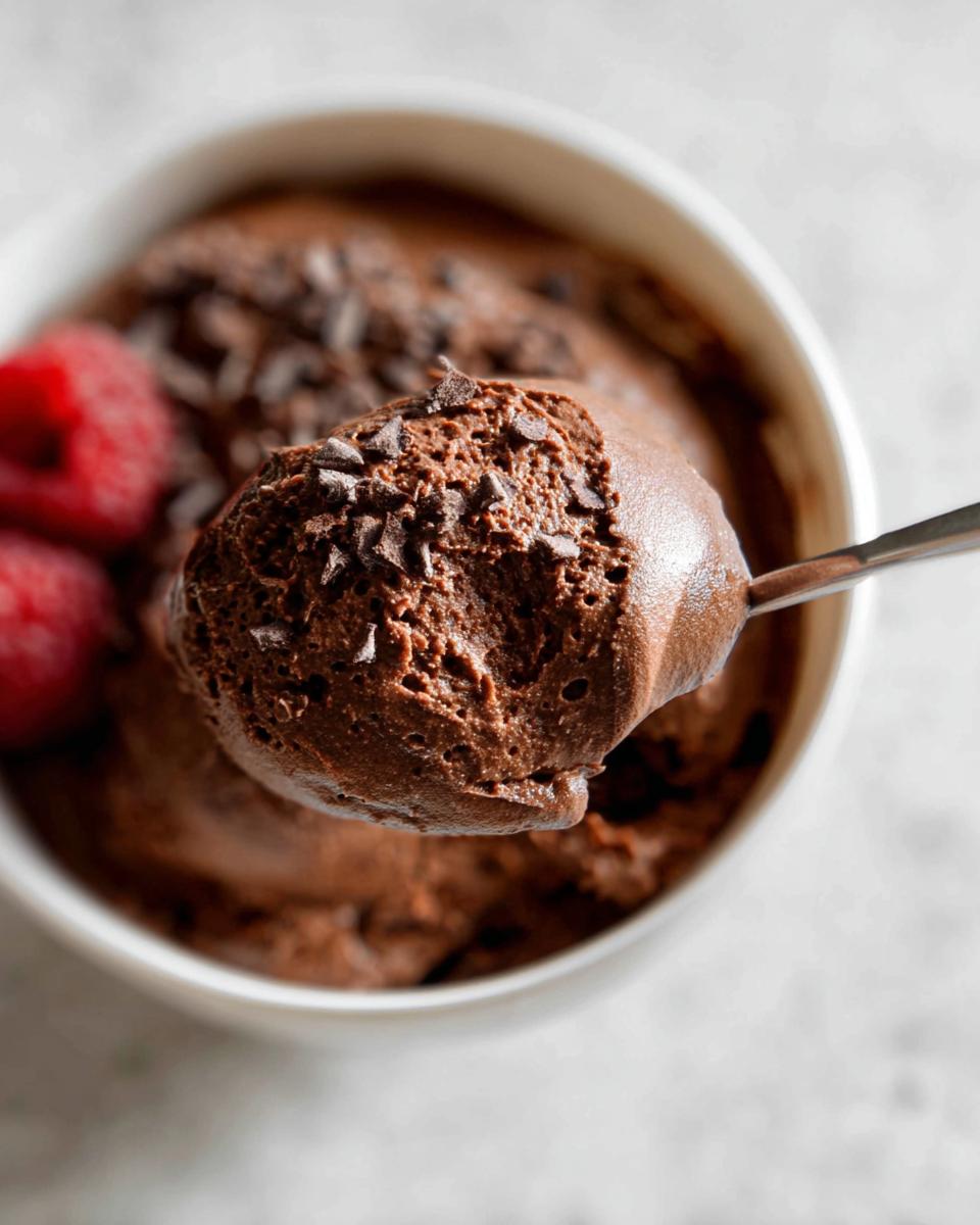 A spoonful of rich High Protein Chocolate Pudding topped with chocolate shavings, next to fresh raspberries in a white bowl.