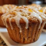 Close-up of a moist peach muffin topped with a drizzled vanilla glaze and chunks of peach.