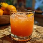 A refreshing peach iced tea slush cocktail in a glass with crushed ice and an orange peel garnish.